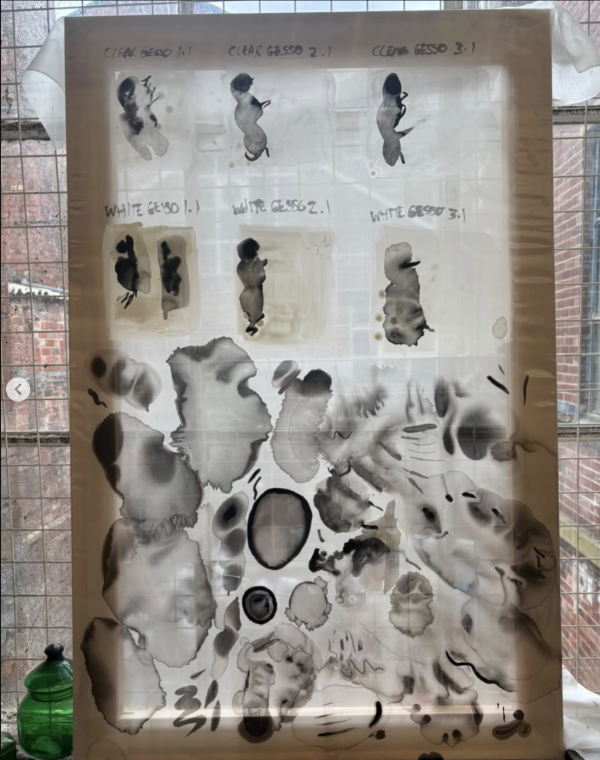 A canvas stretched with silk leans against a window. Test splotches of black ink cover the canvas.