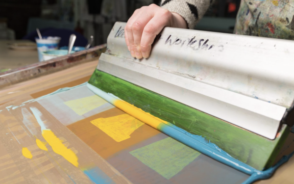 A squeegee being used to screen print an image of plant pots in shades of blue and yellow ink