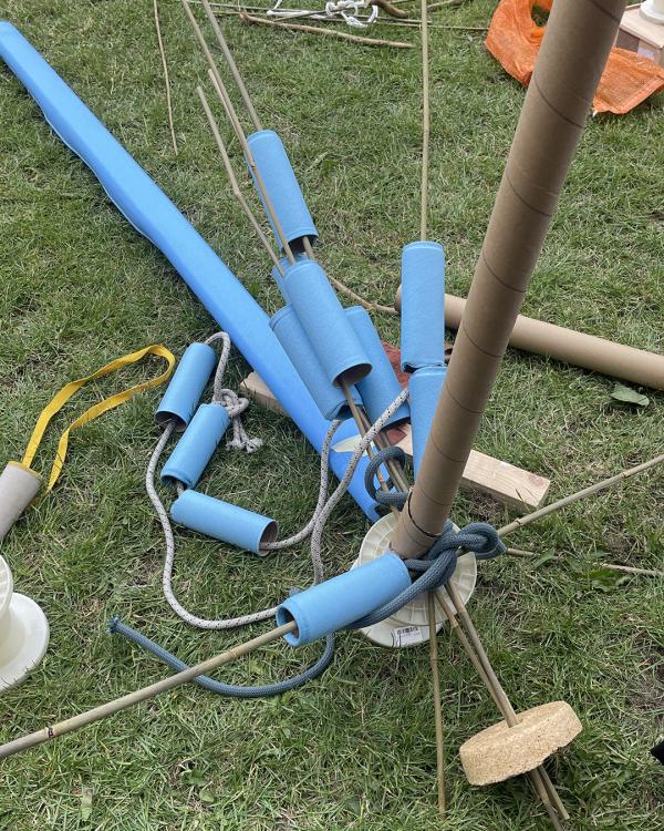 Assorted blue foam tubes, ropes, and sticks are scattered on grass, creating a playful and chaotic assembly. The scene conveys creativity and experimentation.