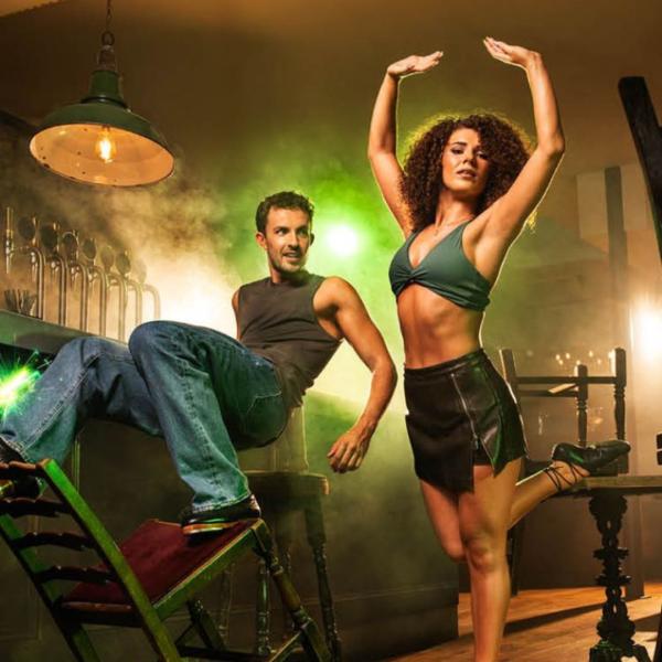 Adam Garcia balances on a chair at a bar, wearing a vest and jeans, looking at another dancer who poses alongside him. A green smoke emits from the corner of the bar. 
