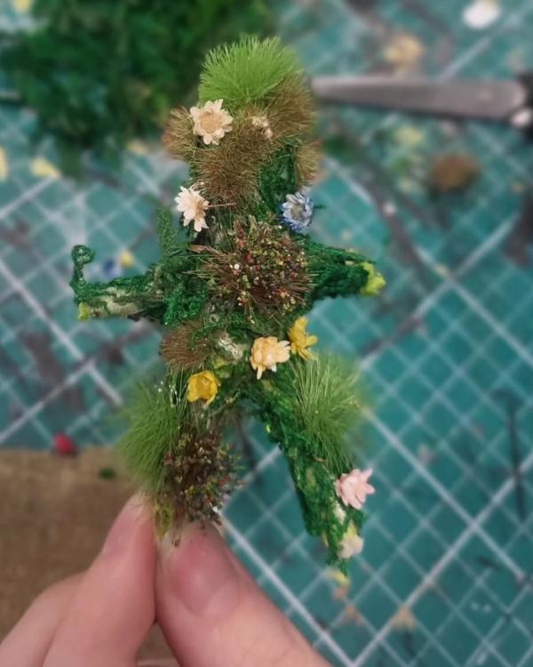 A corn dolly made from recycled materials including fake grass and flowers and held up by a finger and thumb