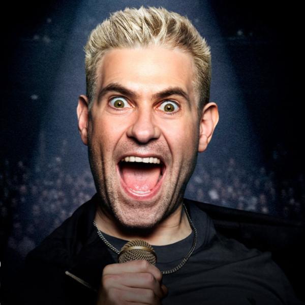 Under a spotlight, Simon Brodkin with a manic expression, wide-eyed and open-mouthed as he holds a microphone up to his chest.