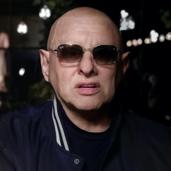 Shaun Ryder holds a neutral gaze, donning a pair of Ray Bans and navy jacket. The background is blurred but busy with lights and activity.