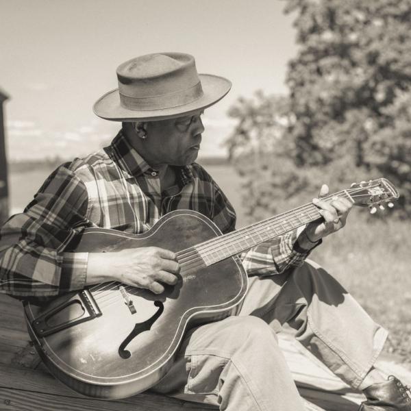 Black and white of Eric Bibb sat outdoors playing a guitar, wearing a plaid shirt and hat shading his face.