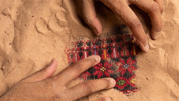 Two hands covered in sand are uncovering a piece of red fabric woven with flowers and a black and red repeating geometric pattern which is buried in the sand. 