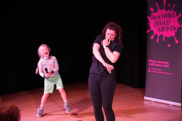 An 7 year old boy with a microphone laughs with delight as a woman strikes a funny pose on a stage