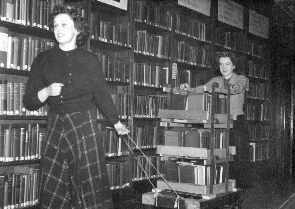 Black and white photo of two librarians moving a large amount of books on a trolley while smiling