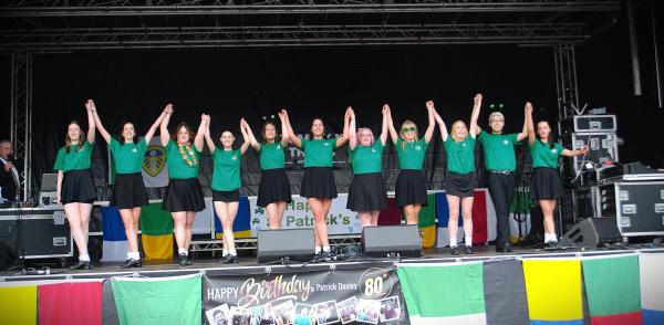 A line of performers on an outdoor stage raise their joined hands, wearing matching green tops and dark skirts or shorts. Flags and banners hang along the front of the stage, with sound equipment and a black backdrop behind them.