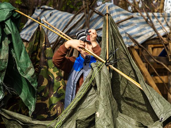 A small child in a striped hat constructing a makeshift shelter with tarps and bamboo poles. They appear focused, surrounded by a woodland setting.