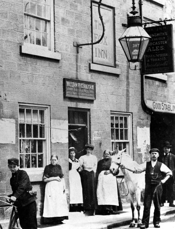 Historic black-and-white photograph of people standing outside the Fox Inn. A horse is held beside the entrance, with a cyclist in the foreground and pub signage visible on the stone building.