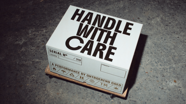 A white delivery box rests on a crate. The box has text on the top of it that reads 'HANDLE WITH CARE'. 