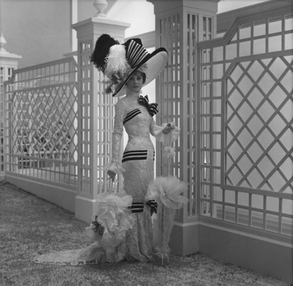 a black and white photograph of a woman in front of a fence, with an ornate black and white striped dress on and large hat with feathers