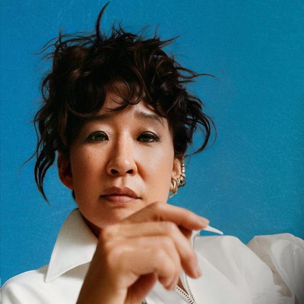 In front of a blue background, a woman with a messy up-do and white shirt looks at the camera, with a hand casually raised in front of her.