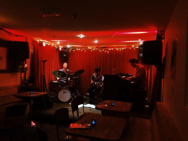 A guitarist, drummer and pianist play on a red lit stage against a red curtain.