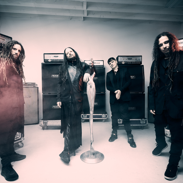 Four members of a rock band Korn dressed in black stand in a studio in front of stacked amplifiers, with a sculptural microphone stand in the centre.