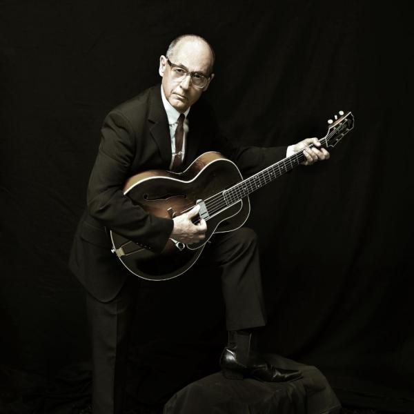 Andy Fairweather Low poses dramatically in a suit, holding an acoustic guitar against a dark, black backdrop.