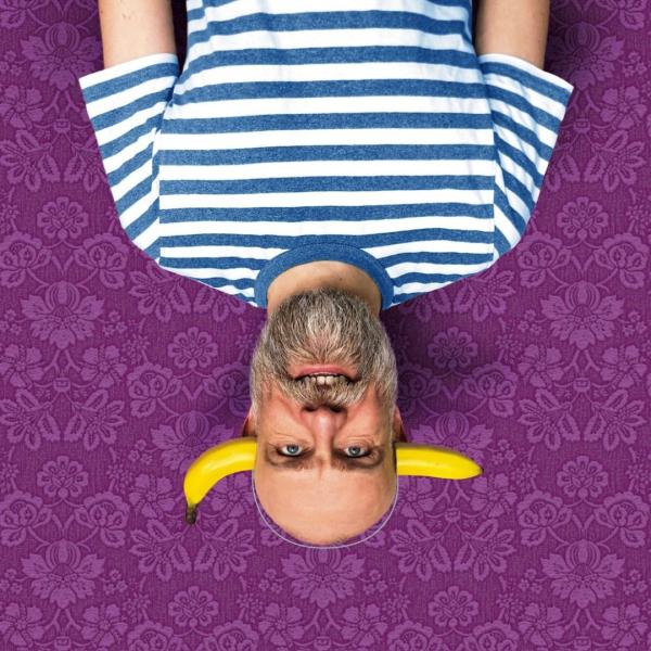 Upside down, Olaf Falafel grins at the camera, wearing a prop banana headband and stripy blue and white t-shirt, positioned over a patterned purple background.