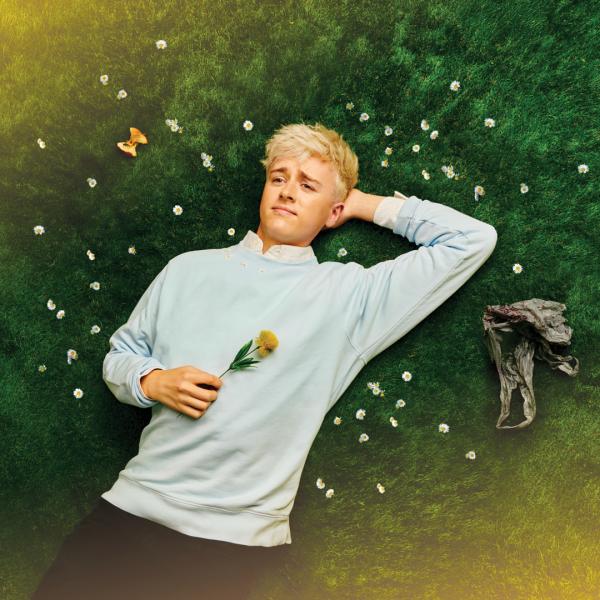 Daniel Foxx lies on a bed of soft grass and gazes at the sky, surrounded by daisies and holding a flower.