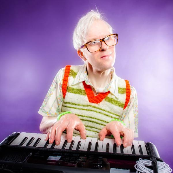 Robert White in glasses and a sweater vest over a patterned shirt, sat at a keyboard in front of a purple backdrop.