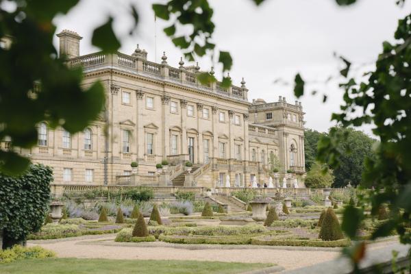Image description: Harewood House from the South front viewed through leafy tree branches, overlooking formal Terrace Garden with clipped hedges, topiary, stone urns, and gravel paths on an overcast day.