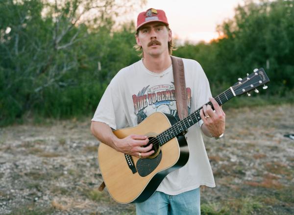 A man wearing a red cap and graphic t-shirt stands outdoors playing an acoustic guitar, with trees and grass in the background.