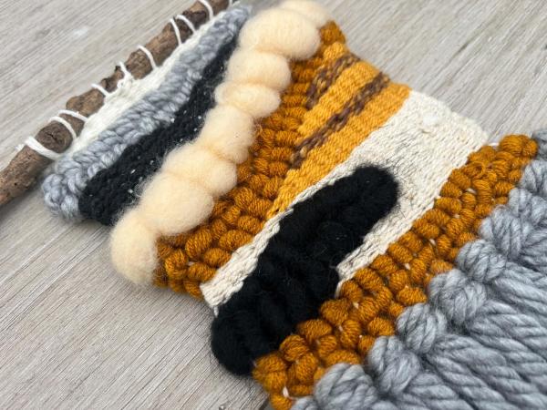 A woven wall hanging in orange, white, black and grey, created with different weaving techniques, lay on a table