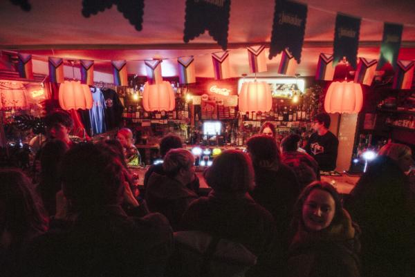 A busy bar with a queue of people waiting, red lights dangle above the bar.