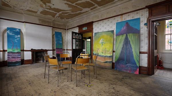 Room with wooden chairs and colorful abstract paintings. Peeling ceiling and walls suggest an aged interior.