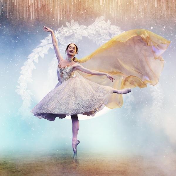 A ballet dancer poses on a pointed toe in a flowy sparkling dress, with a silky yellow cape trailing in the air behind her. Snow, smoke and icicles fill the backdrop.