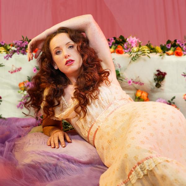 Catherine Bohart lounges on purple fabric in a long floral dress, surrounded by flowers in the background.