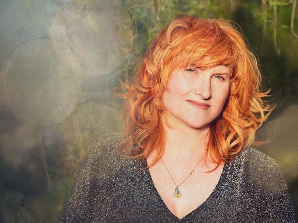 A woman with long vibrant ginger hair and a necklace smiling at the camera.