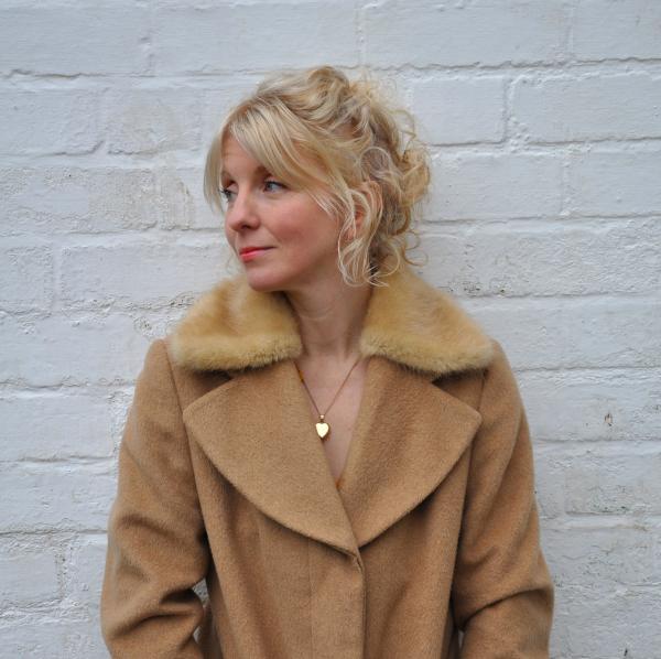 A blonde woman in a beige coat looks off camera in front of a white wall.