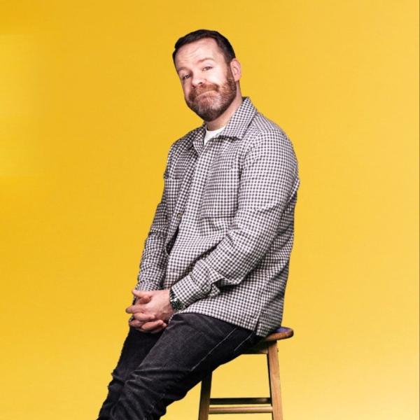 Neil Delamere perches on a high wooden stool in front of a yellow background. 