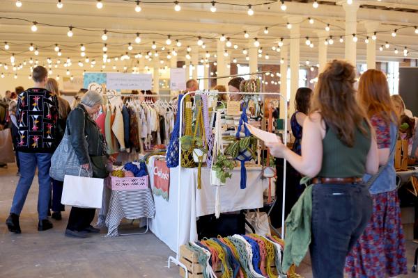 Visitors walking around a busy textile market space at last year's Threads Festival. The venue is an industrial former textile mill with red brick ceilings, and festoon lights hanging from the beams.