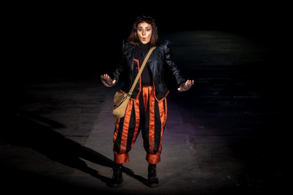 A person wearing a leather jacket and stripped orange trousers stands on a dark stage pulling a surprised face.