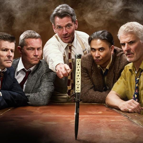 Five men in suits sit together in a smoke-filled room, all looking at a knife in the middle of a table. 
