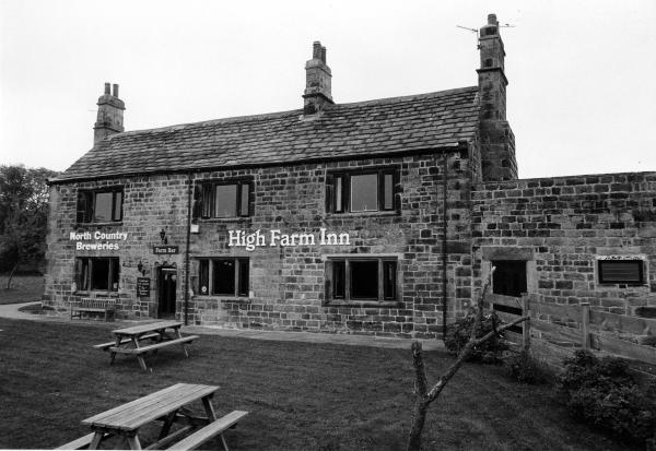 Front view of the stone-built High Farm Inn, displaying signs for North Country Breweries and Farm Bar, with picnic benches on the lawn outside.