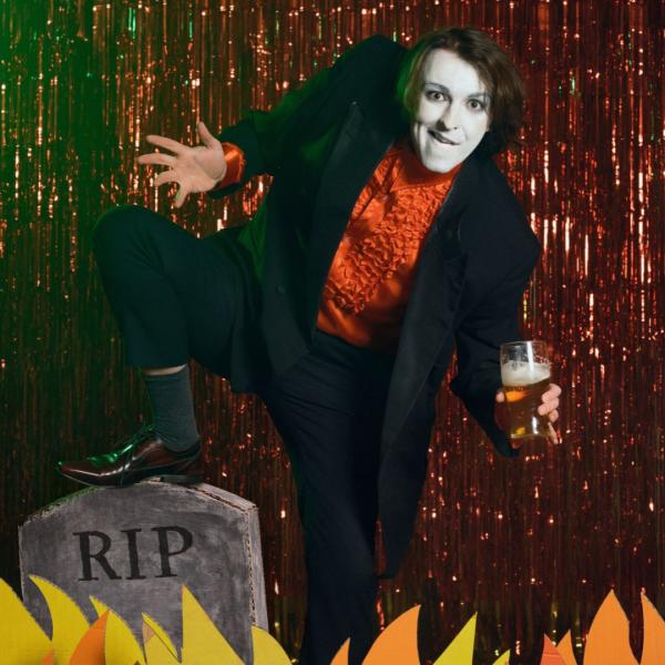 In white face paint and a frilly red shirt, Frankie Monroe grins and holds a pint of beer in front of a sparkly, red stage curtain, his foot rests on a prop gravestone amidst a row of cardboard flames.