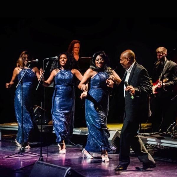 Three singers in sequined blue dresses dance by standing microphones with Roy Hemmings.