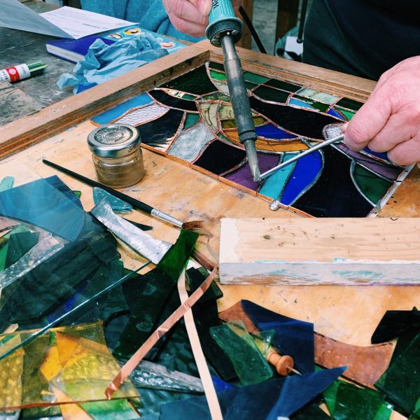 A photograph of someone using a soldering iron to melt solder onto a stained glass piece. There are shards of different coloured glass in the foreground.