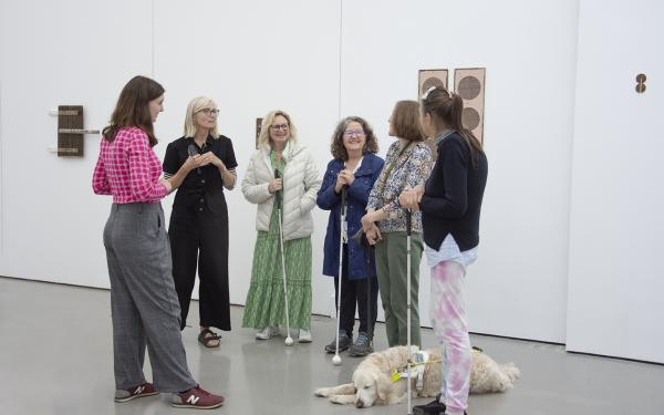 Six people standing in a gallery. they are talking and smiling. Three have white canes, one is holding a magnifying glass, and another has a guide dog resting at her feet.