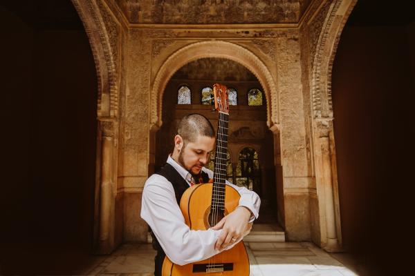 Male figure in formal wear holding an acoustic guitar, the background is a historic building