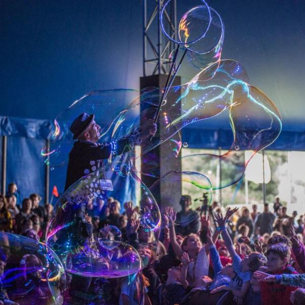 A man in a pork pie hat and polka dot waistcoat creates large bubbles on-stage in a festival tent, as children and families reach out in the audience.
