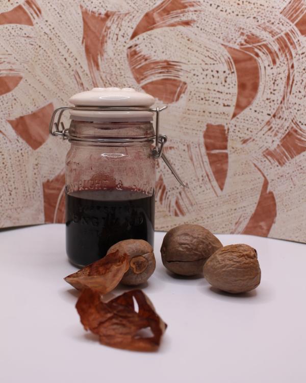 A glass jar filled with avocado ink, sealed with a white lid, placed beside three large avocado stones and their skin, against a roughly painted piece of paper.