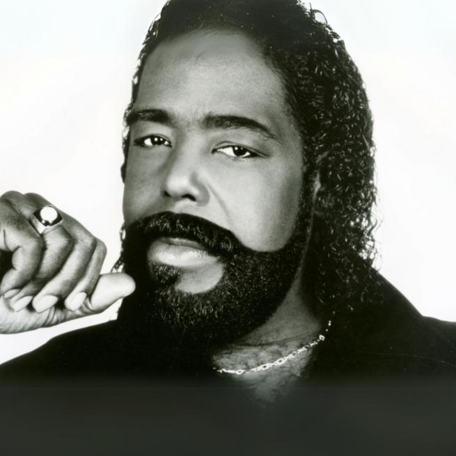 Barry White photographed in black and white, set against a blank backdrop. He has a tight perm and carefully shaped beard, wearing a ring and chain necklace.