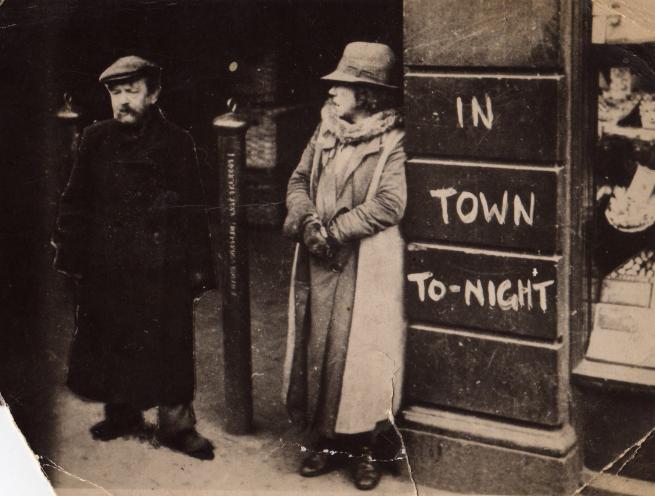 Historic black-and-white street photograph showing two people standing beside a building entrance. Painted text on the wall reads “IN TOWN TO-NIGHT”.