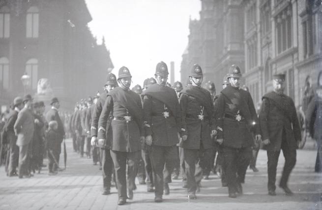 Black and white picture of Victorian policemen marching in formation while pedestrians look on. To the left is Leeds Town Hall. Picture taken in the Leeds Gas Strike of 1890.