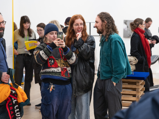 A group of people stood surrounded by art in a white walled gallery. They are smiling and one is taking photos on a mobile phone.
