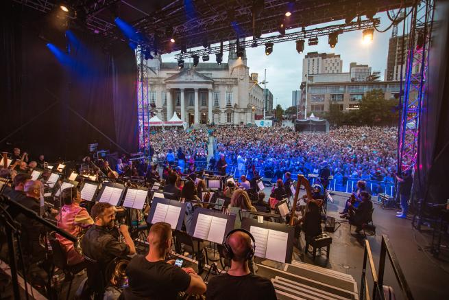 The orchestra of Opera North performing on the Millennium Square stage in front of thousands of people. 