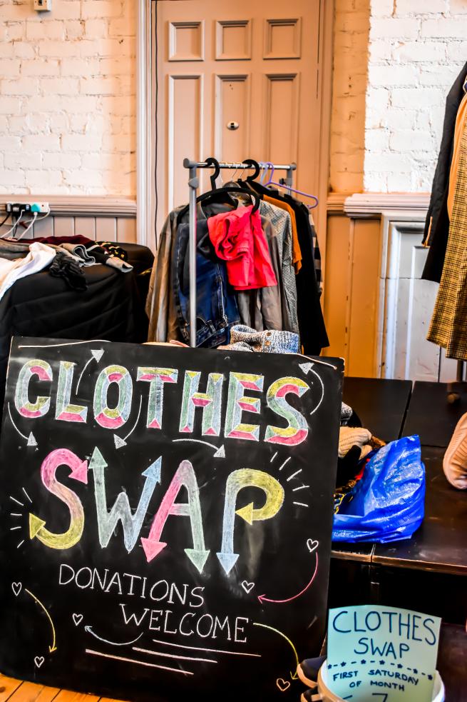 A chalkboard wih the words 'Clothes Swap, Donations welcome' written on in coloured chalk. Behind the sign are rails of clothes.